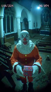 A bald, unnaturally tall figure in a tattered crimson clown suit, face painted in cracked white greasepaint, with a grin stretched too wide—lips split at the corners, revealing blackened teeth. His eyes are wide, bloodshot, and too still. In his gloved hand, he carries a small, wrapped box tied with frayed red ribbon. He doesn’t run. Doesn’t laugh. He walks slowly, deliberately, toward the camera mounted above the confessional. At 00:47 seconds, he stops. Lifts the gift. Holds it out—as if offer