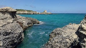 The ruins of Torre di Maradico - a watchtower in Roca Vecchia, located along Italy's southeast coast - overlooks the Adriatic Sea in a daytime view, turquoise water of the area in the foreground.