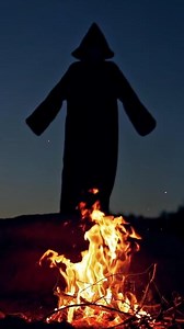 Black death behind the fire flame outdoors. Scary witch in black costume waving arms making fear on the background of burning fire. Halloween costume. Vertical