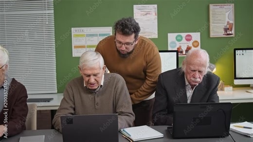 Following front view shot of caring male teacher overseeing group of seniors using laptops during computer literacy class and assisting adult students one by one