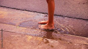 Bare feet of a child stand in a puddle at the base of a driveway. They jump in slow motion and land with a splash. The puddle is reflecting colorful sunset light. Play after the rain. Oil on water too