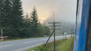 Magical bright sunrise from behind mountain forest through thick white fog looking through open window of moving train. A view through the window of the thick morning fog and the bright sunrays