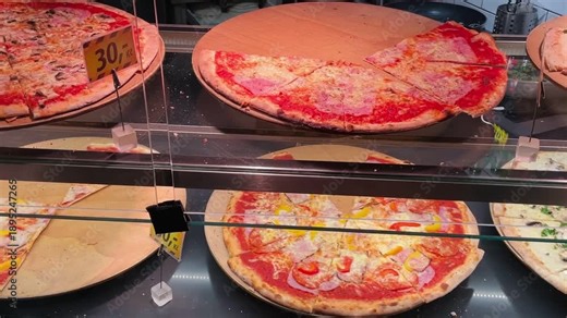 pizza slices in glass display case with warm margherita and herb pizzas on rotating trays, melted mozzarella and bright tomato sauce, golden crispy crusts, Video taken on mobile device.
