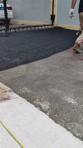 He is applying asphalt coating or liquid tar onto the road surface carefully using a spray tool