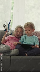 Sister bothers with airplane toy younger brother while watching videos on tablet. Cute children use modern gadget sitting on sofa in living room