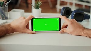 Young adult analyzing horizontal greenscreen display on smartphone sitting at desk, looking at isolated mockup background with copy space. Holding telephone with chroma key. Close up. Tripod shot.