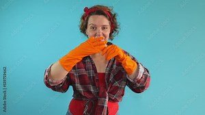 Adult pin-up cleaning woman in a plaid shirt removes orange cleaning gloves from her hands on a blue isolated background. Portrait of woman cleaning. Slow motion portrait