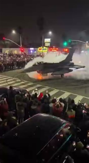 A rooftop cellphone video captured a wildly surreal moment during a late-night street takeover in Los Angeles: a full-size fighter jet spinning donuts in the middle of a city intersection. The F-16-style aircraft roared in a low-power spin as smoke blasted from its wheels and heat waves distorted the neon lights around it. Crowds pressed against the street corners, shouting and filming as the jet swung dangerously close to the curb. Sparks erupted as its wheels carved dark streaks across the pav