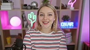 Cheerful young blonde woman streamer triumphs at gaming, flashing thumbs up in joyful success! beaming positively, she's a winner at night in her homely computer room.