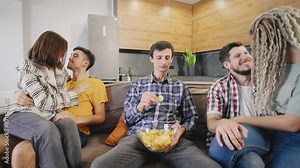 Lonely single man sitting on sofa and eating chips while watching two loving couples of friends in intimate moments, showing dissatisfaction at home party