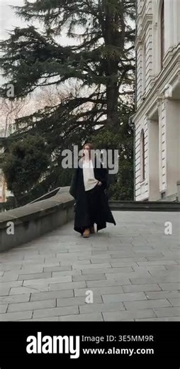 A girl in a coat walks along a stone path with a building and trees in the background during the day Stock Video Footage - Alamy