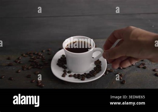 Persons hand reaching for a mug of steaming black coffee on a small plate, uncovering a heart made of coffee beans on a polished wooden table Stock Video Footage - Alamy