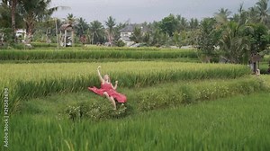 young beautiful girl sits on a green rice field enjoying the nature and beauty around, just arrived on the island, put on a long bright dress and beautifully spread her skirt on the grass. Slowly