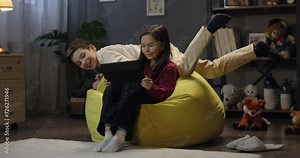 Little girl using digital tablet watching online video cartoon when preteen boy jumps on beanbag. Children using tablet for social media app together in the evening at home