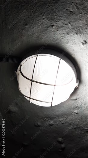 Industrial bulkhead light with a protective cage flickers on a rough concrete ceiling. The erratic illumination creates a harsh, eerie glow in a dark, suspenseful environment.