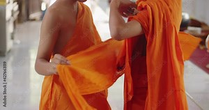 Slow motion scene of the Buddhist novice who has just passed ordination in the summer during school holidays is helping an other novice to dress in a yellow-orange robe.