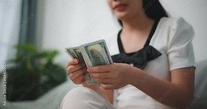 Footage of Happy young asian woman counts cash dollar bills while sitting in living room at home,counting stack cash money dollar