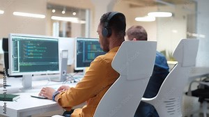 Rear of young man IT programer working on desktop computers typing on keyboard coding a program and listening to music in headphones. Close up. Professional worker writing website in modern office Stock Video