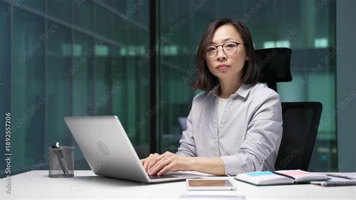 Portrait of serious asian businesswoman sitting at desk at workplace in modern business office. Confident female employee in glasses and casual shirt working on laptop computer and looking at camera