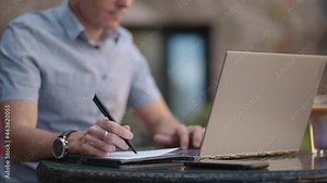 A man sitting in a street cafe works remotely writing a pen in a notebook. Work at the laptop. A man works remotely on a laptop