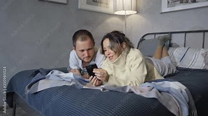 Young couple in hotel bed read texts on smartphone and laugh