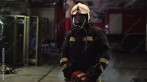 Firefighter portrait wearing full equipment, oxygen mask, and chain saw. Smoke and fire trucks in the background.