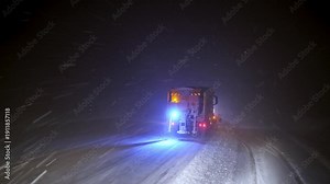 View from inside a vehicle following a snowplow clearing a snow-covered road at night. Blowing snow, flashing warning lights and low visibility show dangerous winter driving and road maintenance opera