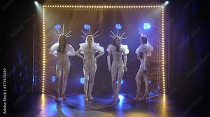 A modern choreographic dance performed by a group of slender young women in tight-fitting flounced overalls and crowns. Shot in a dark studio with neon lights in the background. Slow motion.