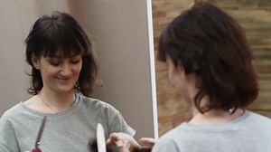 Pretty brunette woman cuts off hair curl with scissors while standing in front of a mirror and smiling. Concept of haircuts and style changes.