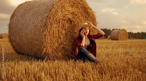Farmer woman in rubber boot sitting at round straw bale and talking by mobile phone on harvested wheat field. Business in agriculture. Farmer in field in rubber boots. Round bale, fertile soil
