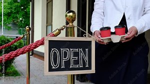 Woman's hands holds two cups of coffee near dashboard with text open. Worker shows advertising opening of the cafe or restaurant.