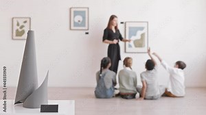 Racking focus from art object on pedestal to children sitting on floor of museum, listening to Caucasian woman talking about paintings on wall