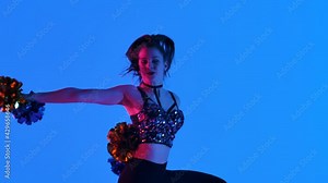 Energetic cheering dance performed by cute cheerleader with pompoms in hands against blue background, illuminated with pink light. Cheerleading, cheering sports team. Close up. Slow motion.
