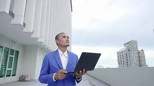 A businessman in a blue suit holds a laptop and looks upward while standing outside a modern building. The scene suggests forward-thinking, technology, and corporate ambition. Stock Video
