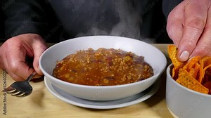 Cinemagraph – Close POV video of steam rising from a bowl of hot chilli con carne, combined with a still image of a man’s hands holding a fork and a tortilla chip from a side dish.