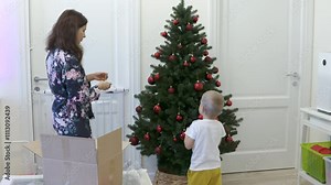 Mother and little boy decorating Christmas tree at home. Happy three year old child helps mom by hanging red ball on christmas tree.