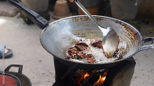 A person stirring meat in a frying pan over an open fire, showcasing traditional outdoor cooking with sizzling oil and visible flames.