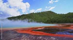A striking geothermal hot spring in Yellowstone National Park radiates with vivid colors as steam rises against a dynamic cloudy sky, showcasing nature's stunning palette. Slow Motion, 4K RAW.