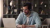 Focused man wearing headphones using laptop writing notes studying...