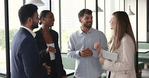 Diverse team of young business colleagues chatting in office hall. Multiethnic office friends discussing corporate life on work break, standing together, talking, smiling, laughing