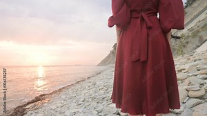 Close-up slow motion behind the legs of a barefoot girl standing in a red dress fluttering in the wind at sunset crouching along the stones of the sea shore. Light Moments of Tidelessness