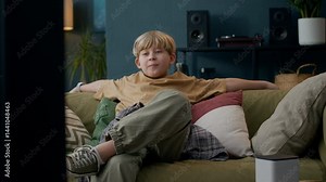 Relaxed Caucasian boy lounging on comfy couch in front of tv set, listening, after interacting with smart speaker, enjoying modern, connected home environment