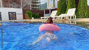 Little girl in swimming pool on funny inflatable donut float ring, learning how to swim. Summer vacation concept. Child floating in a hotel outdoor pool.