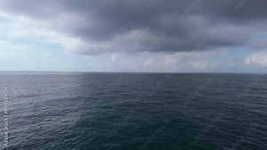 Slow-motion drone flight over the open ocean. Waves ripple under a cloudy sky with sunlight peeking through. The horizon blends sky and water.
