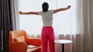 Rear back view woman standing in front of window stretch neck muscles turn head to sides raise hands up