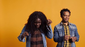 Lovely cheerful husband and wife dancing around in the studio and having fun together, acting silly with groovy funny dance moves against yellow background. Funky married people. Camera B.