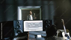 Dolly-in shot of young male singer in glasses and jeans jacket standing in isolation booth of recording studio and singing into microphone while sound engineer using mixing desk in control room