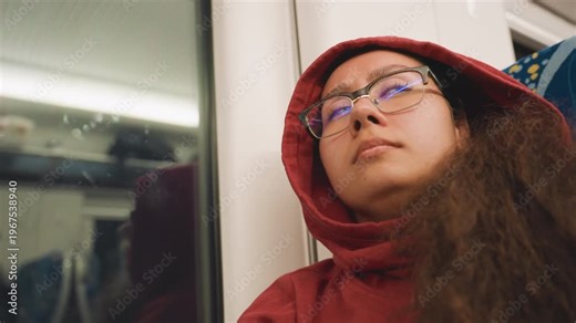 Young artist dozing beside train window, overnight transit with neon reflections, red hoodie framing face, glasses catching city lights, head tilted toward glass, soft bokeh, introspective atmosphere