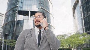 Camera moving around positive Caucasian male. Man talking actively on smartphone while looking around. Man surrounded by large buildings with many glass windows. Searching for correct address.