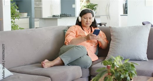Mature Asian woman looking at blue phone tapping scrolling listening via white headset at home